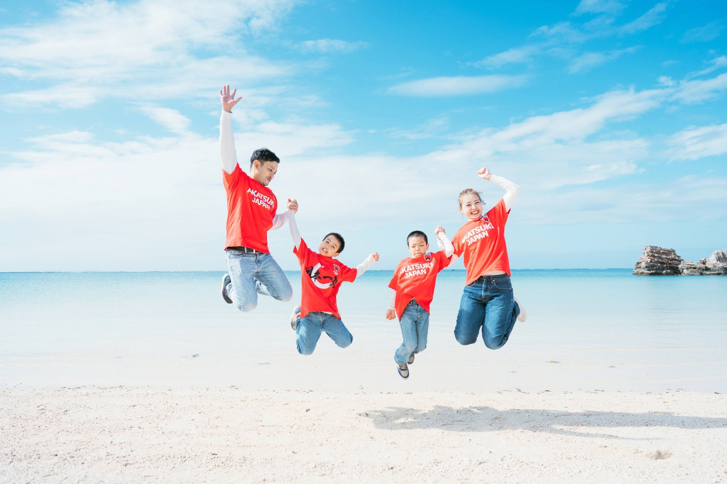 青い空と海を背景に、家族4人でジャンプする撮影写真